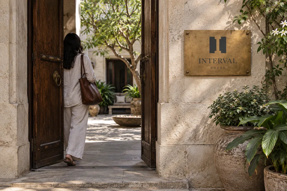 A guest enters Interval House through a stone-and-timber doorway beside an aged brass brand plaque and planted courtyard.