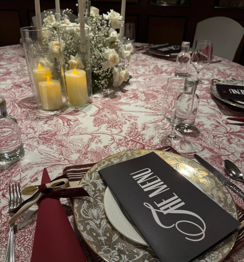Photograph of a dinner table dressed in burgundy toile, with white flowers, candles, gold-rimmed plates and black menu covers.